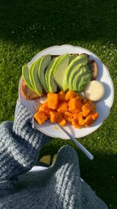 Plate of avocado toast with sweet potatoes held over green grass in sunlight.